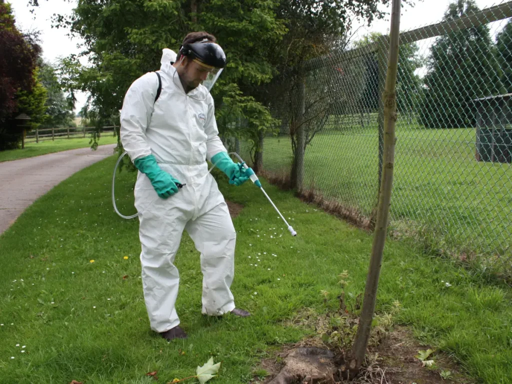 A professional applicator in full Personal Protective Equipment applying to the base of a tree using a Micron AccuDos sprayer